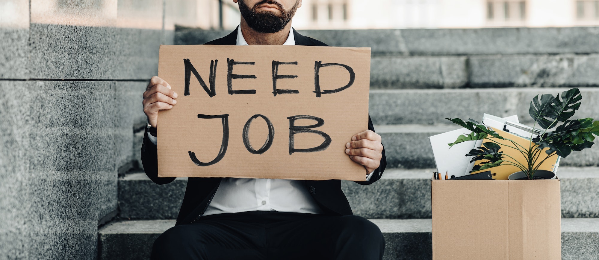 Unemployment concept. Mature businessman sitting on stairs with poster Need Job and box of personal